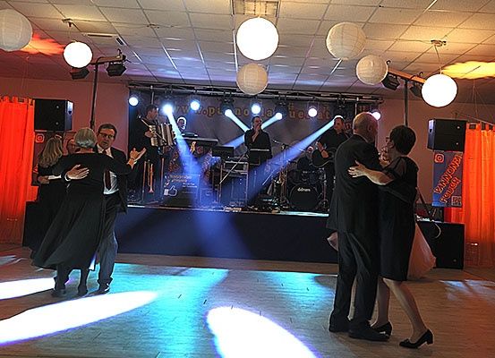 Mehrere Paare tanzen in einem Ballsaal mit einer Bühne hinter ihnen, auf der eine Band mit Mikrofon und Instrumenten steht. Die Bühne ist mit blauen und orangefarbenen Lichtern beleuchtet.