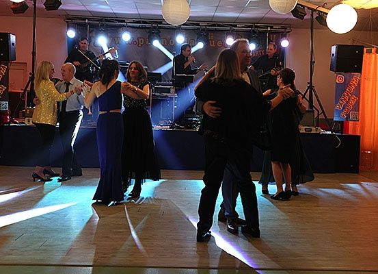 Eine Gruppe von Menschen tanzt auf einer Bühne unter Scheinwerfern, mit einer Band, die hinter ihnen spielt, und Lautsprechern auf beiden Seiten.