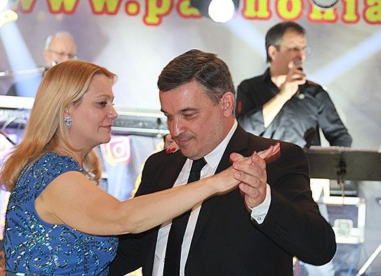 Ein Mann und eine Frau tanzen auf der Bühne, während ein Musiker hinter ihnen spielt. Die Frau trägt ein blaues Kleid und der Mann einen Anzug und eine Krawatte. Ein Banner im Hintergrund zeigt ein Logo.