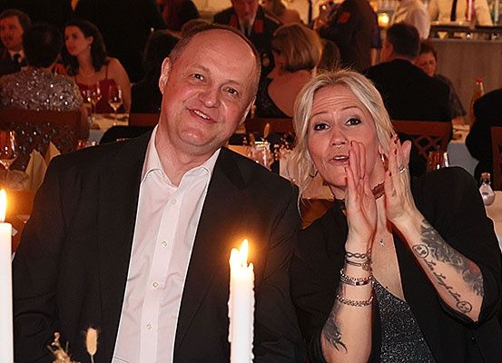 Ein Mann und eine Frau sitzen bei brennenden Kerzen an einem Tisch, umgeben von anderen Gästen. Die Frau hat Tattoos am Arm.
