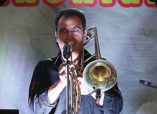 Ein Mann mit Brille spielt eine Posaune auf der Bühne und trägt ein schwarzes Hemd. Hinter ihm ist ein weißer Hintergrund mit rotem und gelbem Text.