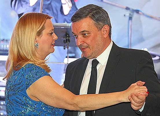 Ein Mann und eine Frau tanzen auf der Bühne, der Mann in einem schwarzen Anzug und einer Krawatte, die Frau in einem blauen Kleid, mit Musikinstrumenten und einem unscharfen Hintergrund.