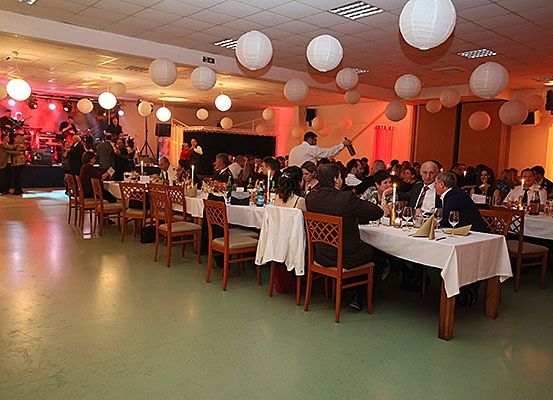 Ein Bankettsaal mit runden Tischen für eine Dinnerparty. Die Leute sitzen um die Tische herum. Der Raum ist mit hängenden Papierlaternen und Vorhängen dekoriert. Es gibt eine Bühne mit einer Band, die im Hintergrund auftritt.
