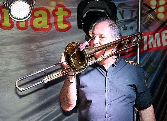 Ein Mann in einem blauen Hemd spielt Posaune auf der Bühne, beleuchtet von einem Scheinwerfer, mit einem Banner, das 'Blat' und 'ZEIT' im Hintergrund liest.