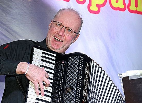 Ein älterer Mann mit Brille spielt ein schwarzes Akkordeon. Er lächelt und scheint aufzutreten. Das Akkordeon hat den Schriftzug 'BELTUNA'. Hinter ihm ist ein weißer Hintergrund mit roten Buchstaben.