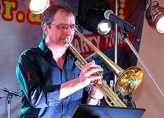 Ein Mann spielt Posaune auf der Bühne vor einem Mikrofon. Er trägt eine Brille und ein blaues Hemd.