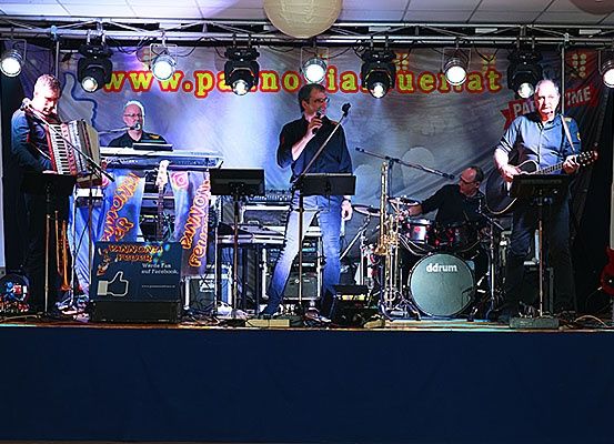 Eine Band spielt auf einer Bühne mit einem Banner, das 'www.panonia.at' besagt. Der Mann in der Mitte singt und hält ein Mikrofon.