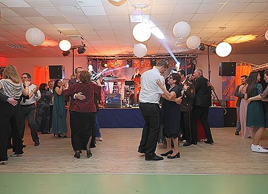 Mehrere Menschen tanzen auf einem Holzboden in einem Raum mit roten Wänden und einer Bühne, auf der eine Band spielt. Es hängen weiße Papierlaternen von der Decke.