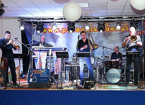 Eine Band spielt auf der Bühne mit einem Banner, Scheinwerfern und Musikinstrumenten. Die Musiker spielen Trompeten, ein Akkordeon und Schlagzeug.