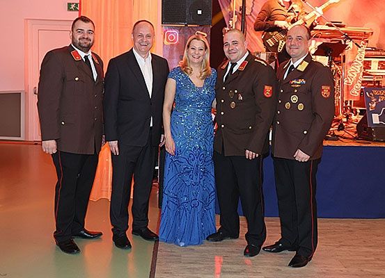 Fünf Personen stehen in formeller Kleidung zusammen; drei Männer in Militäruniformen und eine Frau in einem blauen Kleid. Dahinter ist eine Bühne mit Musikinstrumenten und einem Künstler aufgebaut.