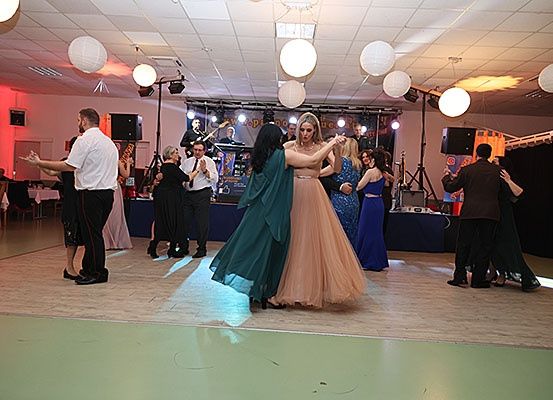 Eine lebhafte Tanzparty mit Paaren, die in einem Saal unter Deckenbeleuchtung tanzen. Eine Band spielt auf der Bühne, und Menschen sitzen an Tischen.
