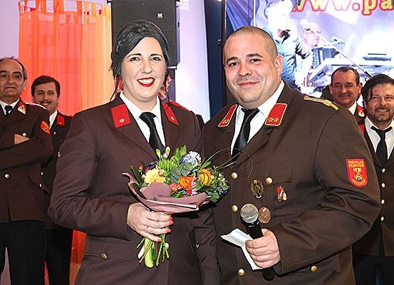 Zwei uniformierte Personen, ein Mann und eine Frau, stehen bei einer Veranstaltung eng zusammen, lächeln und halten einen Blumenstrauß und ein Mikrofon. Hinter ihnen steht eine größere Gruppe uniformierter Personen, einige lächeln, vor einem Banner.