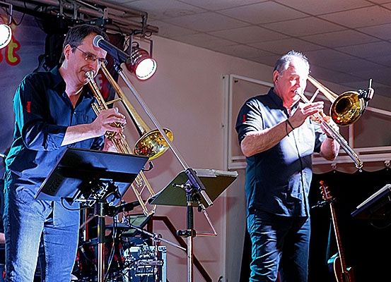Zwei Männer spielen auf der Bühne mit Posaunen, einer von ihnen spielt auch Trompete. Vor ihnen steht ein Mikrofon. Dahinter befindet sich ein Schlagzeug und eine Gitarre. Eine Lampe hängt an der Decke.