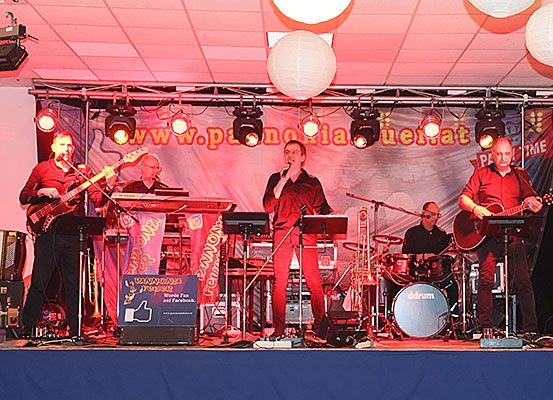 Eine Band tritt auf einer Bühne mit roter Beleuchtung auf. Der Leadsänger singt und hält ein Mikrofon. Die Bandmitglieder spielen verschiedene Instrumente. Auf der rechten Seite der Bühne steht ein Schlagzeug.