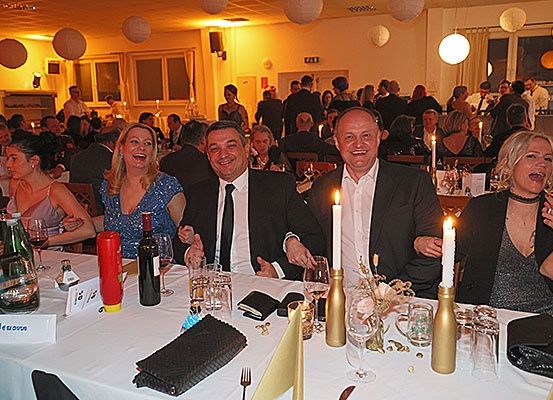 Eine Gruppe von Menschen sitzt an einem Dinnertisch mit Kerzen, lächelt und posiert für ein Foto. Flaschen, Gläser und andere Gegenstände stehen auf dem Tisch. Der Raum hat warmes Licht und Papierlaternen hängen von der Decke.