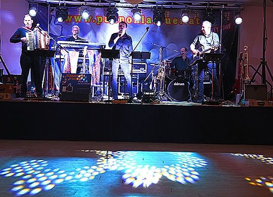 Eine Band tritt auf einer Bühne mit einer Projektionsleinwand auf. Ein Mann singt in ein Mikrofon, und ein anderer spielt Akkordeon. Die Bühne ist von Lichtern beleuchtet, und der Boden hat eine blumenartige Projektion.