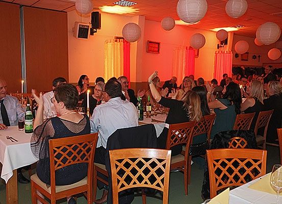 Eine Gruppe von Menschen sitzt um Tische in einem Bankettsaal herum, wahrscheinlich um ein Ereignis zu feiern. Flaschen, Weingläser und Servietten sind auf den Tischen. Einige Leute stehen und klatschen. Der Raum ist mit hängenden Laternen und Vorhängen dekoriert.