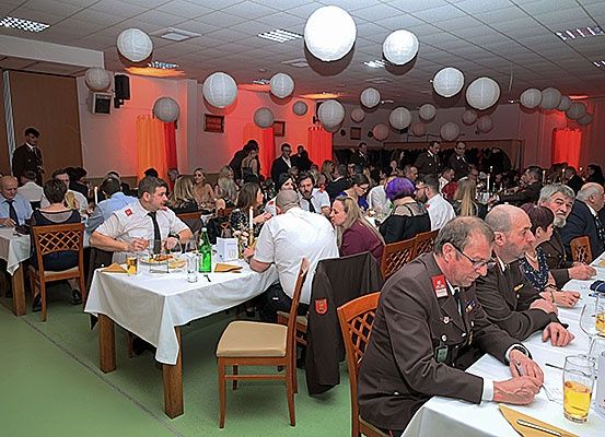 Eine formelle Veranstaltung findet in einem Bankettsaal mit Tischen und Stühlen statt. Menschen in formeller Kleidung sitzen, einige mit Brille und Uniform. Sie trinken aus Gläsern und essen von Tellern. Es hängen Papierlaternen an der Decke.