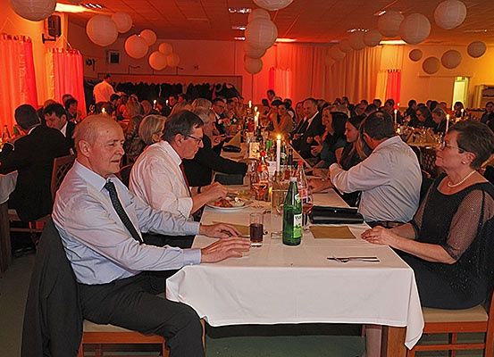Eine Gruppe von Menschen sitzt in einem Bankettsaal an Tischen, in formeller Kleidung und in Gesprächen vertieft. Die Tische sind mit Tellern, Weingläsern und Kerzen geschmückt. Der Raum ist mit roten Vorhängen und Papierlaternen, die von der Decke hängen, dekoriert.