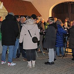Eine Gruppe von Menschen in Winterkleidung steht nachts vor einem Gebäude, einige mit Getränken, in einer festlichen Atmosphäre.