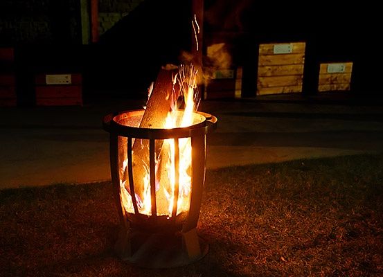 Ein Feuerkorb mit einem brennenden Holzstamm in der Nacht. Ein großer Metallrahmen umgibt das Feuer. Hinter dem Feuer sind Holzlagerboxen zu sehen.