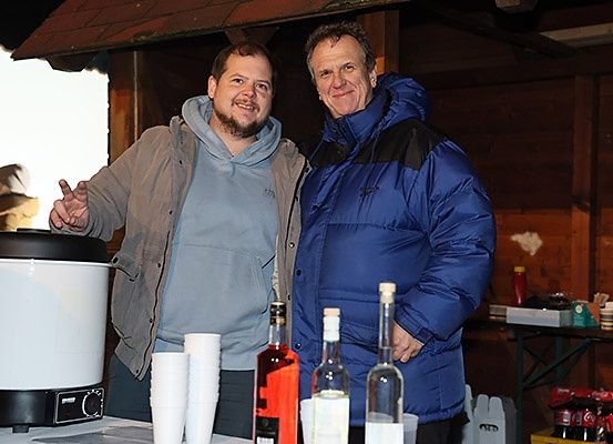 Zwei Männer stehen neben einem Tisch mit Flaschen und Bechern. Einer zeigt mit dem Finger und lächelt, der andere lächelt und trägt eine blaue Jacke. Hinter ihnen steht ein großes weißes Gerät und ein Tisch mit verschiedenen Gegenständen.