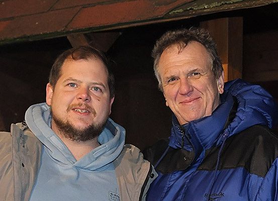 Zwei Männer stehen eng beieinander und lächeln. Einer trägt eine blaue Jacke, der andere eine blau-schwarze Jacke. Sie scheinen für ein Foto zu posieren.