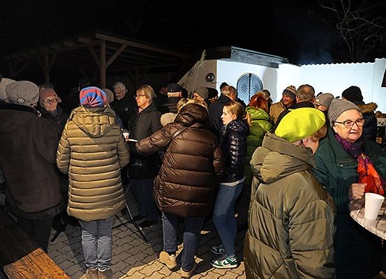 In der Nacht stehen eine Gruppe von Menschen in warmer Kleidung um ein Gebäude, einige halten Tassen. Eine Frau trägt einen bunten Hut, und eine andere hält eine Kamera. In der Nähe ist eine Bank, und im Hintergrund befindet sich ein Feuerkorb.