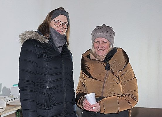 Zwei Frauen stehen in einem Raum. Eine hält einen Becher, die andere schaut in die Kamera. Beide tragen warme Kleidung, eine hat eine Kapuze mit Pelzbesatz. Hinter ihnen steht ein Tisch mit Gegenständen.