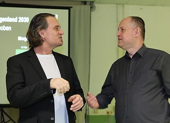 Zwei Männer stehen vor einer Projektionsleinwand und diskutieren möglicherweise etwas. Der Mann auf der linken Seite trägt einen schwarzen Anzug.