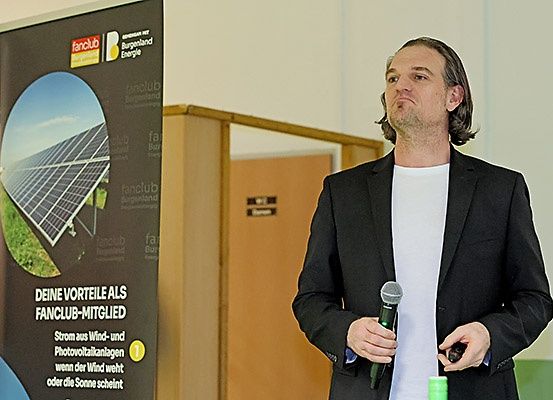 Ein Mann in einem schwarzen Anzug hält ein Mikrofon in einem Konferenzraum, mit einem Plakat mit Solarmodulen dahinter.
