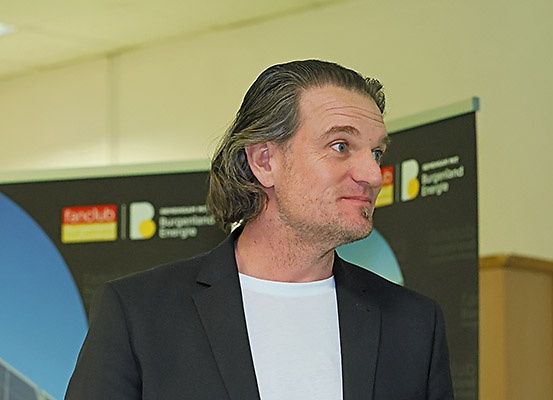Ein Mann mit langen Haaren und Bart, in einem schwarzen Anzug und einem weißen T-Shirt, steht drinnen, wahrscheinlich in einem Konferenzsaal. Hinter ihm ist ein großes Banner mit Logos und Text zu sehen.