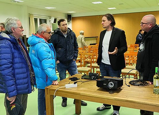 Fünf Männer stehen um einen Holztisch mit einem Projektor und Kabeln herum. Sie befinden sich in einem Raum mit Holzwänden und einem grünen Boden.