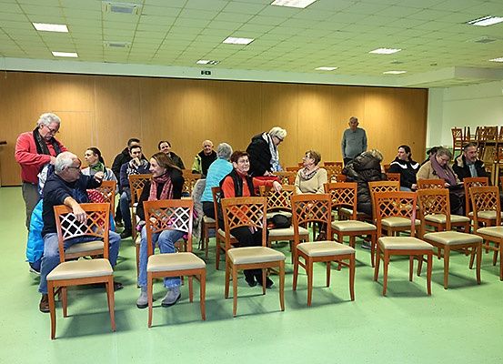 Eine Gruppe von Menschen sitzt in einem Raum mit Holzstühlen. Der Raum hat Holzwände und grünen Boden. Ein Mann steht vor ihnen, und eine Lautsprecher ist an der Wand montiert.