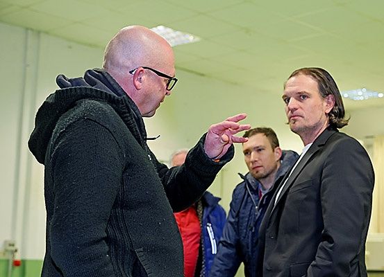 Ein Mann mit Brille gestikuliert, während er mit einem Mann im Anzug spricht, mit zwei anderen Männern in Jacken hinter ihnen in einer Innenumgebung.