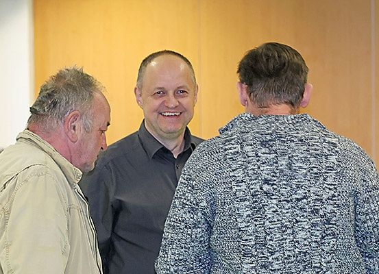 Drei Männer stehen in einem Raum. Der Mann auf der linken Seite schaut den lächelnden Mann in der Mitte an. Der Mann auf der rechten Seite schaut den Mann in der Mitte an. Sie stehen vor einer Holzwand.