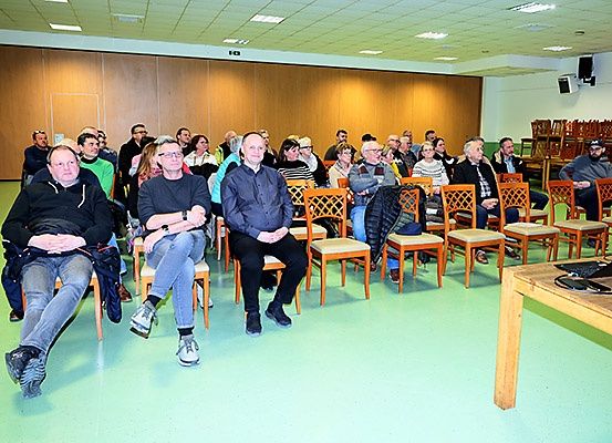 Eine Gruppe von Menschen sitzt in einem Raum, einige mit Brillen und Jacken. Stühle sind in Reihen angeordnet, und ein Holztisch mit Laptop steht rechts.