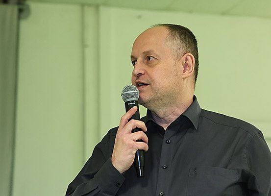 Ein Mann mit Mikrofon steht vor einer grünen Wand und hält vermutlich eine Rede.