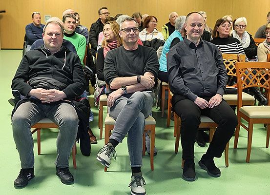 Drei Männer sitzen in einem Raum mit anderen Personen auf Stühlen. Der Mann auf der linken Seite trägt eine schwarze Jacke. Der Mann in der Mitte trägt eine Brille und eine Uhr. Der Mann auf der rechten Seite lächelt.