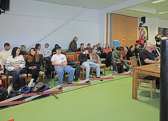 Eine geschlossene Versammlung mit Menschen auf Stühlen, einer Person in der Ecke und einem Holztisch mit einer Flasche darauf.