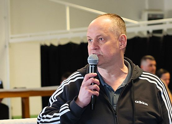 Ein Mann in einer Adidas-Jacke hält ein Mikrofon und spricht zu einer kleinen Menge. Hinter ihm ist eine weitere Person teilweise sichtbar.