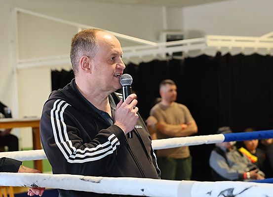 Ein Mann spricht in ein Mikrofon in einem Boxring, während einige Leute hinter den Seilen zusehen.