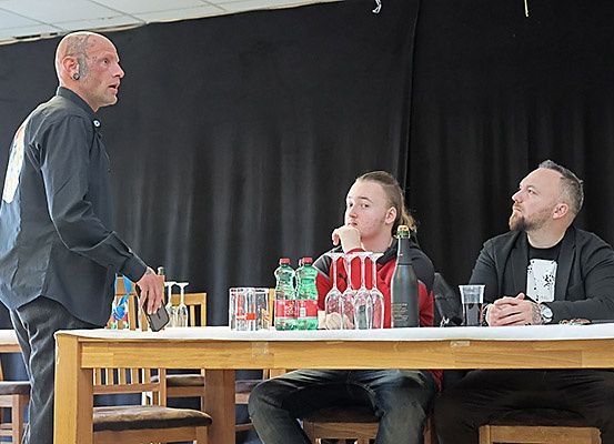 Ein Mann steht neben einem Tisch mit Flaschen und Gläsern, während zwei sitzende Männer zusehen, möglicherweise auf einer Bühne.