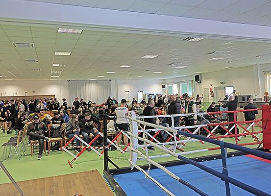 Eine Menschenmenge versammelt sich in einer Boxhalle, um einen Boxkampf zu verfolgen. Zuschauer sitzen auf Stühlen, während andere um den Ring herum stehen, der von roten Seilen umgeben ist.