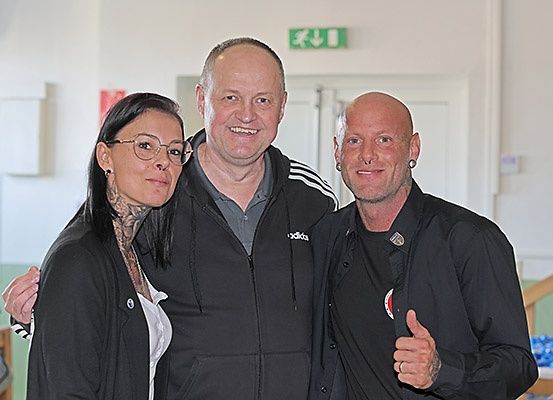 Drei Personen posieren für ein Foto, zwei Männer und eine Frau. Die Frau hat Tattoos am Hals, und die Männer lächeln. Sie befinden sich in einem Raum mit weißen Wänden und einem grünen Fluchtwegschild.