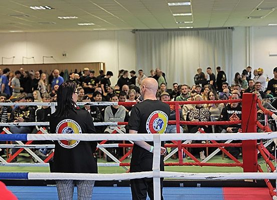 Zwei Personen stehen in einem Boxring und blicken auf eine Menge. Der Ring ist von roten Seilen und blauen Matten umgeben. Das Publikum sitzt in Reihen und beobachtet die Veranstaltung.