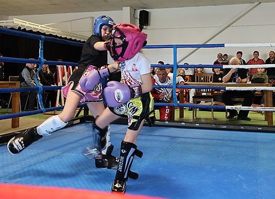Zwei junge Boxer kämpfen in einem Ring mit blauen Matten. Einer trägt ein schwarzes Shirt, der andere ein weißes. Zuschauer beobachten vom Rand aus.