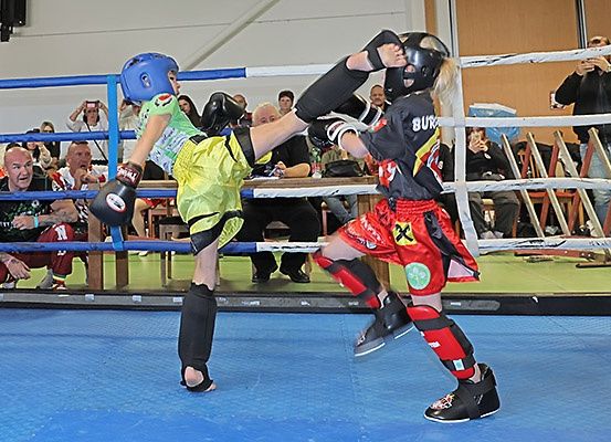 Zwei Kinder boxen in einem Ring, tragen Helme und Handschuhe. Das Publikum schaut von den Seitenlinien zu.