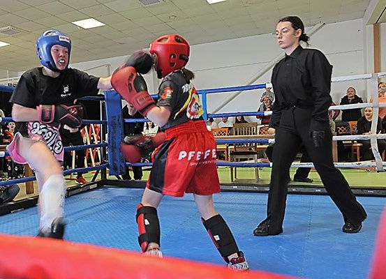 Zwei junge Boxer im Ring, einer in Blau, der andere in Rot, bei einem Kampf, den ein Schiedsrichter beobachtet. Der Ring ist blau und Zuschauer beobachten von den Seitenlinien.