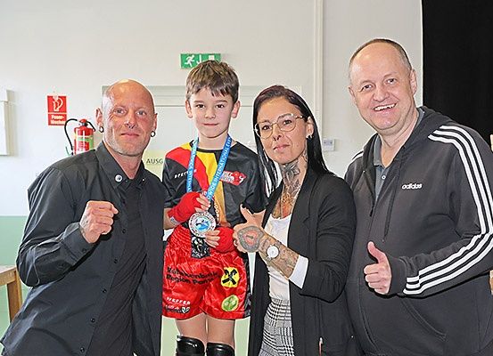 Vier Personen posieren für ein Foto mit einem jungen Jungen in roter Boxausrüstung, der eine Medaille hält.
